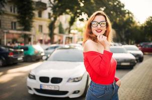 atractiva mujer pelirroja con anteojos, blusa roja y falda de jeans posando en la calle contra un auto deportivo blanco. foto