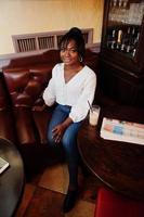 Stylish african american women in white blouse and blue jeans posed at cafe with caramel latte. photo