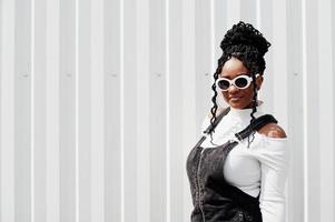 African woman in denim overalls skirt, white sunglasses posed against white steel wall. photo