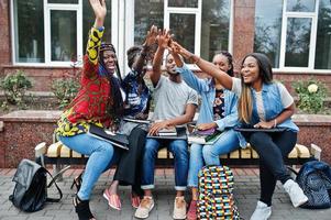 Group of five african college students spending time together on campus at university yard. Black afro friends studying at bench with school items, laptops notebooks. photo