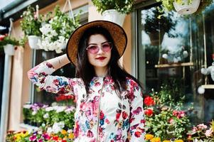 retrato de verano de una chica morena con gafas rosas y sombrero contra la tienda de flores. foto