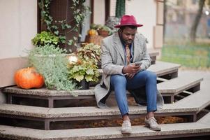 Stylish African American man model in gray coat, jacket tie and red hat sitting against pumpkins. Autumn mood. photo