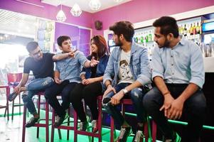 Group of stylish asian friends wear on jeans sitting at chairs against bar in club and chatting together. photo