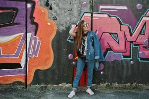 Stylish casual hipster girl in jeans wear and glasses against large graffiti wall. photo