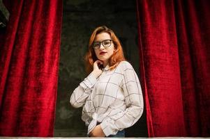 Attractive redhaired woman in eyeglasses, wear on white blouse posing at arch of open red curtains. photo