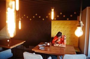 Confident young indian man in orange sweater sitting at pizzeria and eat pizza. photo