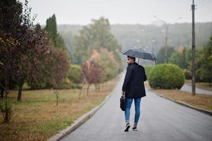 hombre de barba árabe alto de moda vestido con abrigo negro con paraguas y estuche posado en el día de la lluvia. foto