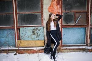 Fashionable long legs brunette model in long black cloak posed outdoor at winter day against old grunge wall with broken windows. photo