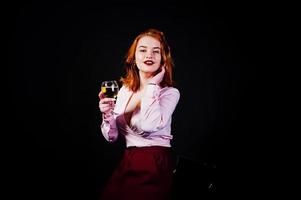 Gorgeous red haired girl in pink blouse and red skirt with glass of wine at hand isolated on black. photo