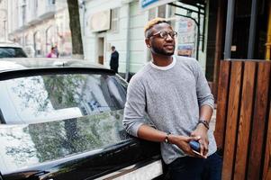 Stylish african american boy on gray sweater and glasses posed at street against black business car. Fashionable black guy. photo