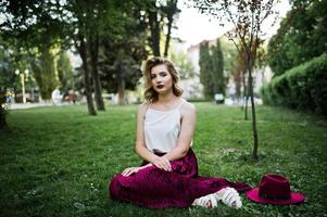Fashionable and beautiful blonde model girl in stylish red velvet velour skirt, white blouse and hat, sitting on green grass at park. photo
