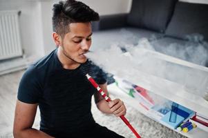 Handsome and fashionable indian man in black sitting at room, smoking hookah photo