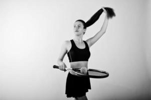 Black and white portrait of beautiful young woman player in sports clothes holding tennis racket while standing against white background. photo