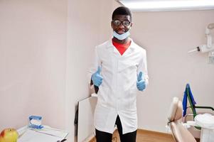 African american male doctor in glasses and mask posed in clinic and shows thumb up sign. photo