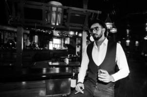 Handsome well-dressed arabian man with glass of whiskey and cigar posed at pub. photo