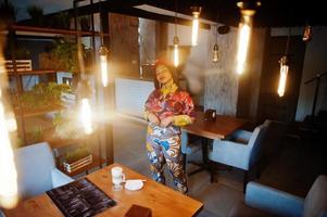 Enthusiastic african american woman in trendy coloured outfit with red beret chilling in cozy cafe with many lamps. photo