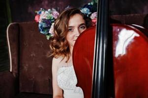 Pretty young gilrl musician in white dress with double bass sitting on brown vintage sofa. photo