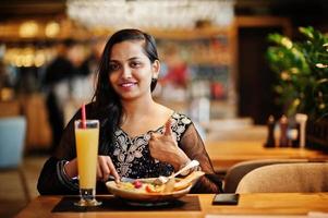 Pretty indian girl in black saree dress posed at restaurant, sitting at table with juice and salad. Show thumb up. photo