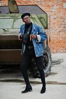 African american man in jeans jacket, beret and eyeglasses, with cigar posed against btr military armored vehicle. photo