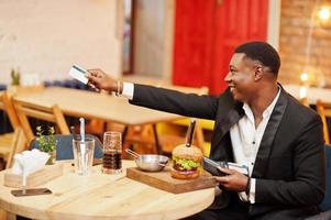 Gives credit card from wallet to paying waiter by food. Respectable young african american man in black suit sitting in restaurant with tasty double burger and soda drink. photo