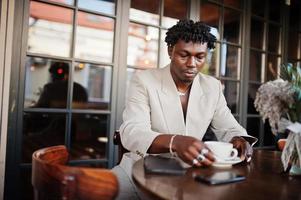 Stylish afro man in beige old school suit sitting on cafe with cup of coffee. Fashionable young African male in casual jacket on bare torso. photo