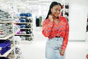 Modish african american woman in  red shirt and jeans skirt posed at clothes store. It's time for shopping. photo