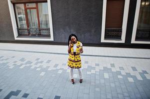 Stylish african american women in yellow jacket posed on street with hot drink in disposable paper cup and mobile phone at hands. photo