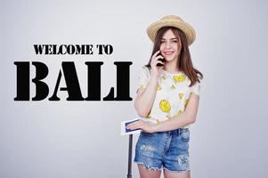 Girl tourist with bag, wear in shirt, shorts and hat, speaking on phone isolated on white. Welcome to Bali. photo