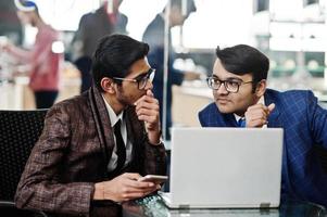 Two indian business man in suits sitting at office on cafe and looking at laptop. photo