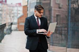 hombre de negocios indio con estilo en ropa formal con portátil en las manos de pie contra las ventanas en el centro de negocios. foto