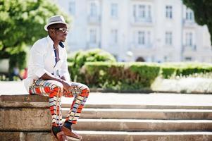 Stylish african american man in white shirt and colored pants with hat and glasses posed outdoor. Black fashionable model boy. photo