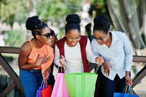 Three casual african american girls with colored shopping bags walking outdoor. Stylish black womans shopping. photo