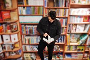 Tall smart arab student man, wear on black jeans jacket and eyeglasses, at library with book at hands. photo