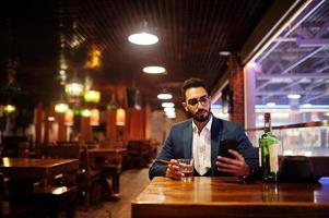 un apuesto hombre árabe bien vestido con un vaso de whisky y un cigarro sostiene un teléfono móvil, posado en el pub. foto