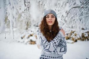 Cute curly girl in sweater and headwear at snowy forest park at winter. photo