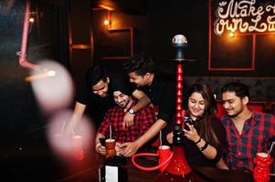 grupo de amigos indios divirtiéndose y descansando en el club nocturno, bebiendo cócteles y fumando narguile, mirando teléfonos móviles. foto