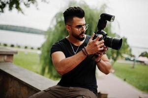 Awesome beautiful tall ararbian beard macho man photographer in glasses and black t-shirt with professional camera at hands. photo