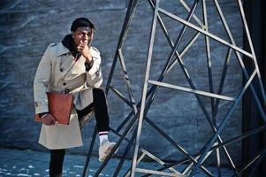 Handsome african american man posing outside in black hat and beige coat with folder in hand. photo