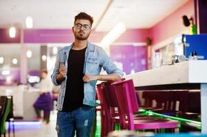 Stylish asian man wear on jeans jacket and glasses posed against bar in club. photo