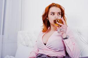 Hermosa chica pelirroja con blusa rosa y falda roja con una copa de vino a mano en la cama de la habitación. foto