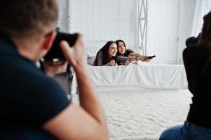 The team of two photographers shooting twins models girls while they click the remote control from the TV on studio. Professional photographer on work. photo