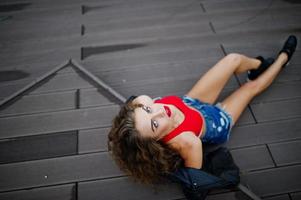 Sexy curly model girl in red top, jeans denim shorts, leather jacket and sneakers posed on the floor. photo