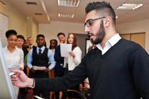 Arabian business coach presenting report standing near whiteboard pointing on sales statistic shown on diagram and chart teach diverse multiracial company members gathered together in conference room. photo