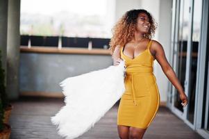 mujer afroamericana glamurosa con vestido amarillo y capa de lana blanca posada en la terraza. foto