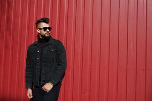 Fashion Arab man wear on black jeans jacket and sunglasses posed against red steel wall background. Stylish and fashionable arabian model guy. photo