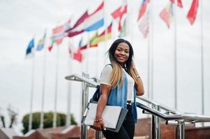 African student female posed with backpack and school items on yard of university, against flags of different countries. photo