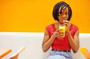 African woman sitting on cafe against orange wall and drink pineapple juice in hands. photo