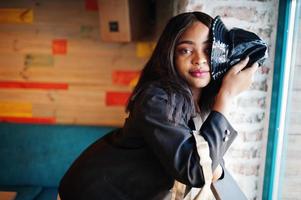 encantadora modelo afroamericana con chaqueta negra y sombrero relajándose en el café durante el tiempo libre. foto