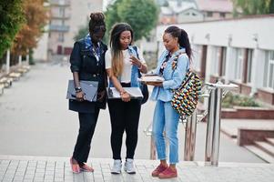 Three african students female posed with backpacks and school items on yard of university and look at tablet. photo