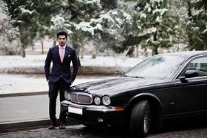 elegante modelo de hombre macho indio con traje y corbata rosa posado contra un coche clásico negro. foto